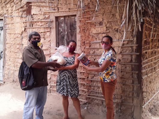 Integrante da ASMUBIP entrega cesta agroecológica para um casal. Eles estão em frente à uma casa de taipa, todos os três utilizam máscara.