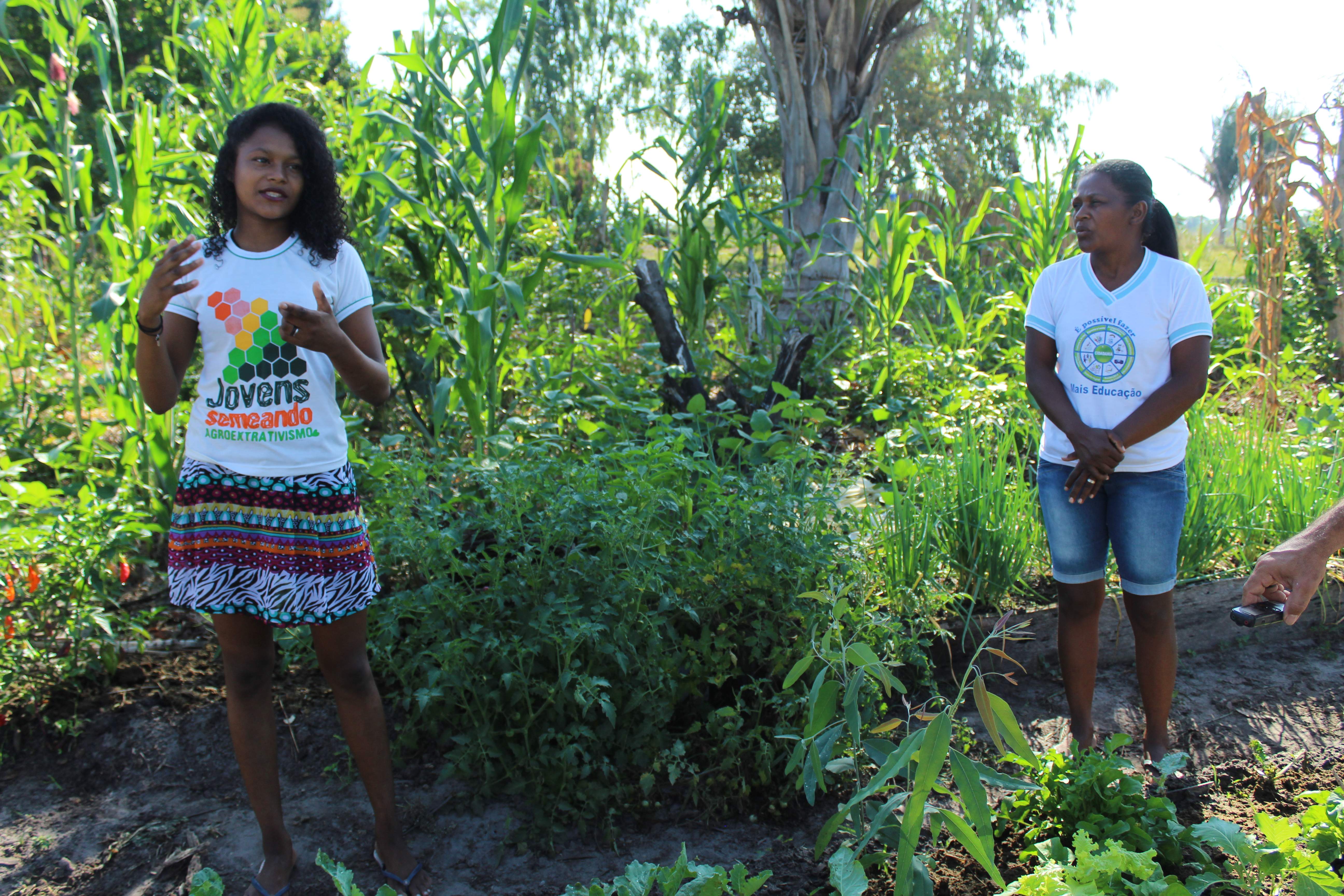 Uma moça está do lado esquerdo da imagem, gesticulando e falando. A mãe da moça está à direita, olhando para ela. Elas duas estão em um ambiente com plantas, como palmeira, milho, verduras em geral 